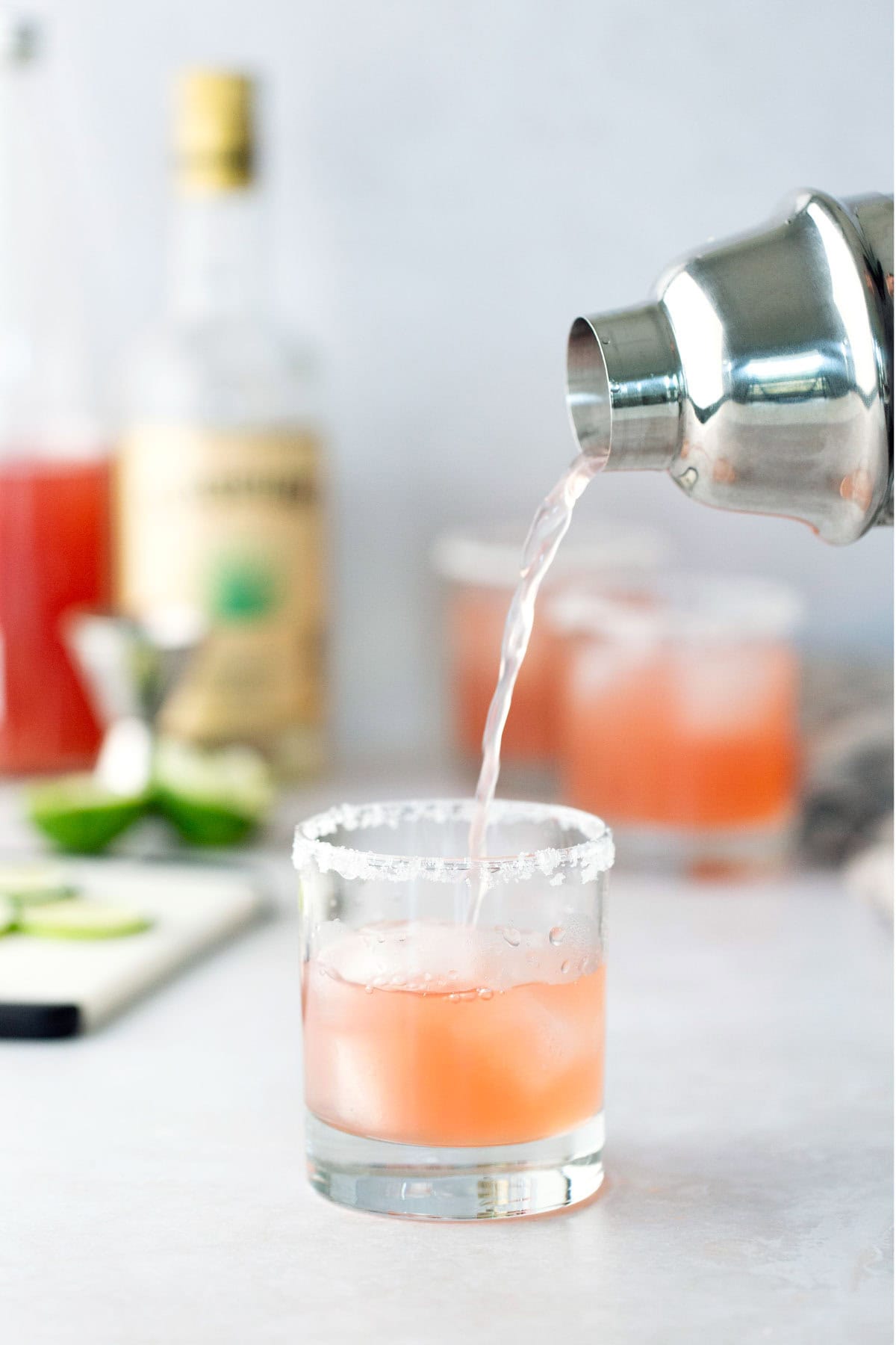 A cocktail shaker pours a pink drink into a salt-rimmed glass with ice; bottles and cocktail ingredients are blurred in the background.