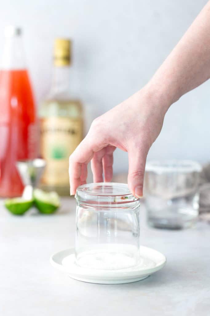 A hand dips the rim of an upside-down glass in a plate of sugar, with bottles and lime wedges blurred in the background.