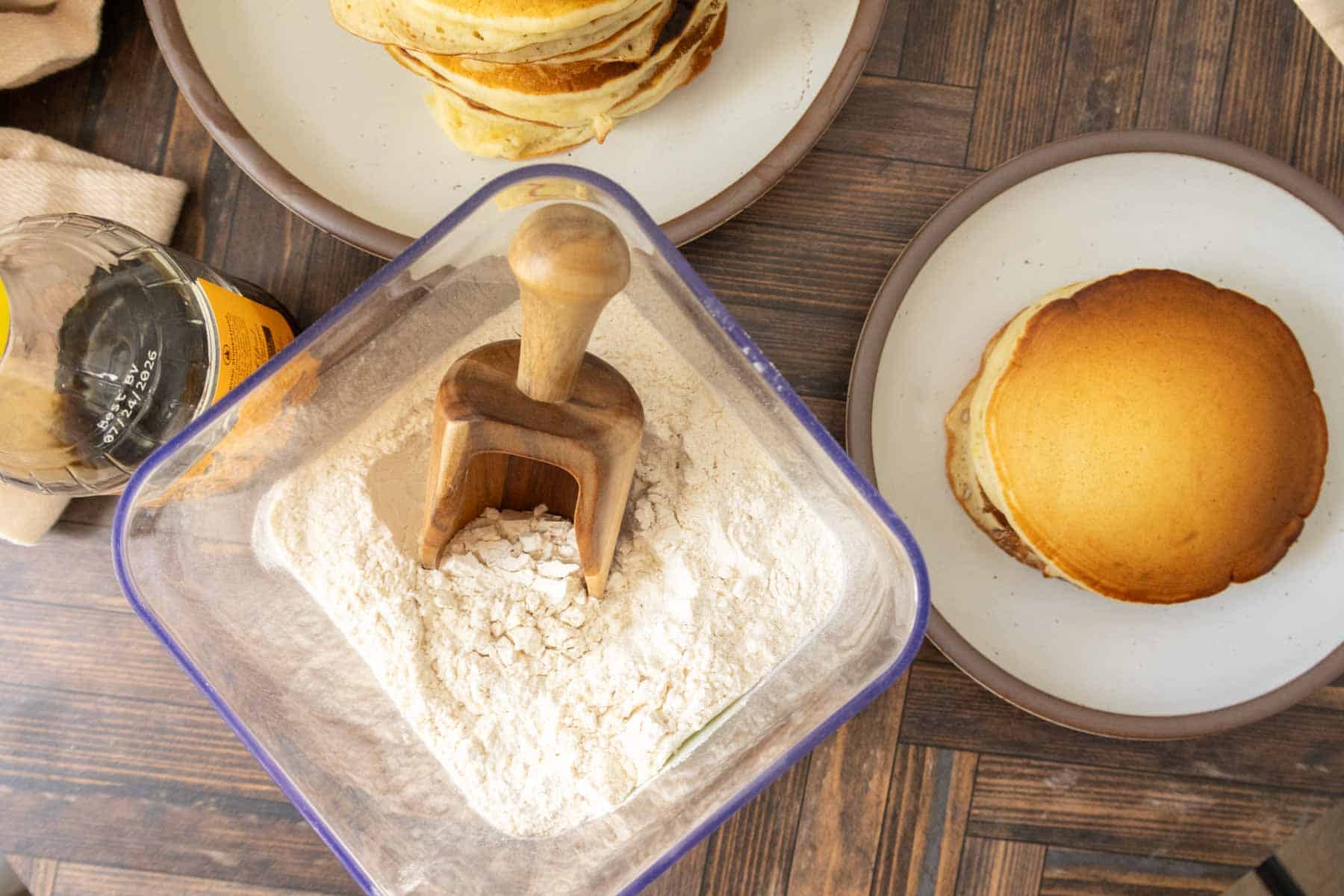 A container of flour with a wooden scoop, a bottle of syrup, and plates of stacked pancakes on a wooden table.
