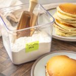 A clear container labeled “Pancake Mix” with a wooden scoop, next to plates stacked with golden pancakes on a wooden table.