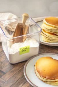 A clear container labeled “Pancake Mix” with a wooden scoop, next to plates stacked with golden pancakes on a wooden table.
