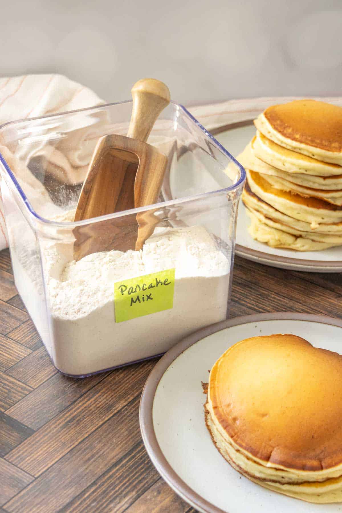 A clear container labeled โPancake Mixโ with a wooden scoop, next to plates stacked with golden pancakes on a wooden table.