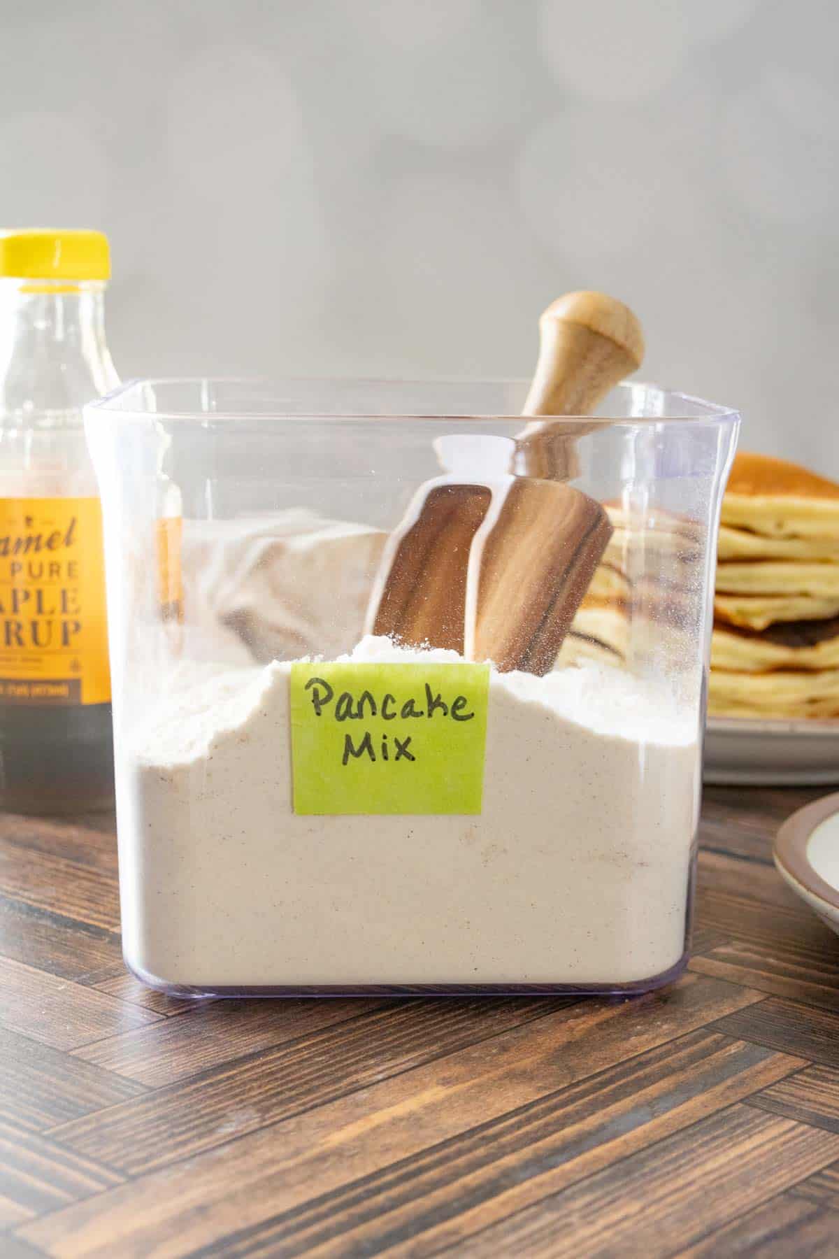 A clear container labeled "Pancake Mix" sits on a wooden surface, filled with powder and a wooden scoop. Syrup and a stack of pancakes are visible in the background.