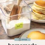A clear container of pancake mix with a scoop and a labeled sticky note sits next to plates of stacked pancakes on a wooden surface.