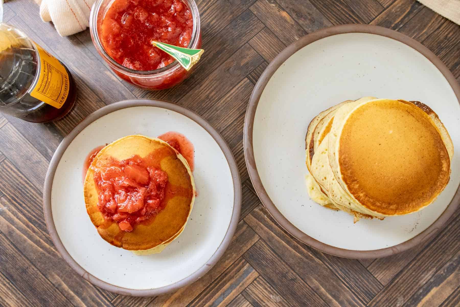 Two plates of pancakes on a wooden table; one plate has plain pancakes, the other has pancakes topped with strawberry sauce. Syrup and a jar of strawberry sauce are nearby.