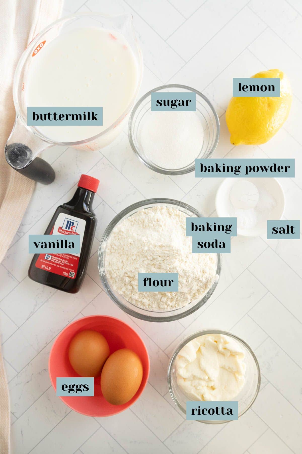 Various baking ingredients are arranged on a white surface, each labeled: buttermilk, sugar, lemon, baking powder, vanilla, flour, baking soda, salt, eggs, and ricotta.