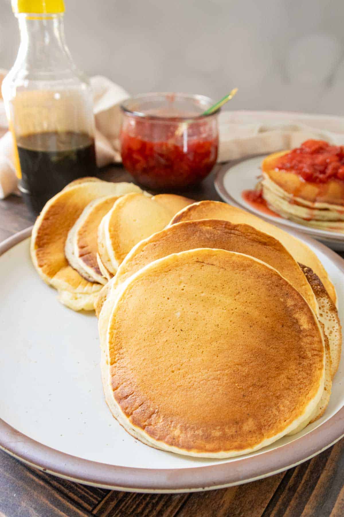 A plate of golden pancakes is in the foreground, with syrup in a bottle and a jar of strawberry jam in the background. Another plate of pancakes with jam is also visible.