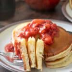 A stack of pancakes topped with strawberry compote, with a fork holding a sliced portion on a ceramic plate.