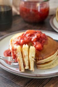 A stack of pancakes topped with strawberry compote, with a fork holding a sliced portion on a ceramic plate.