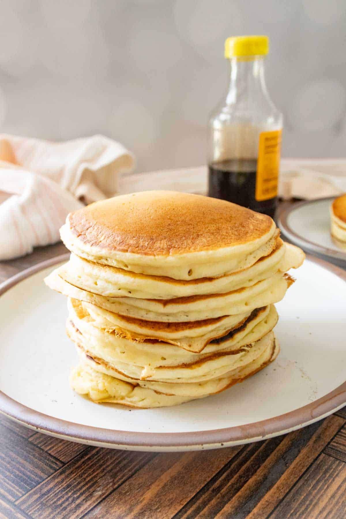 A stack of pancakes sits on a plate with a bottle of syrup in the background on a wooden table.