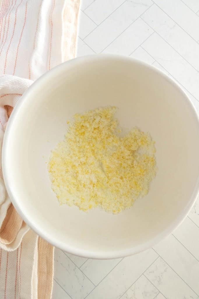 A white mixing bowl with a small amount of a yellow and white granular mixture inside, placed on a light countertop near a striped kitchen towel.