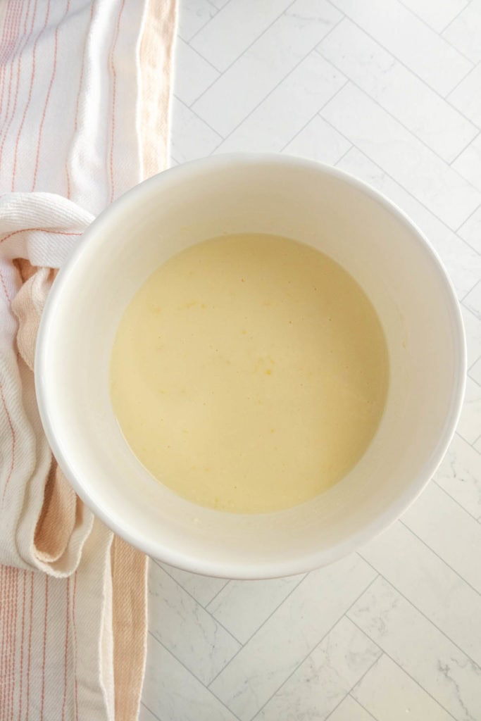 A white mixing bowl with pale yellow batter sits on a light-colored, herringbone-patterned countertop next to a striped kitchen towel.