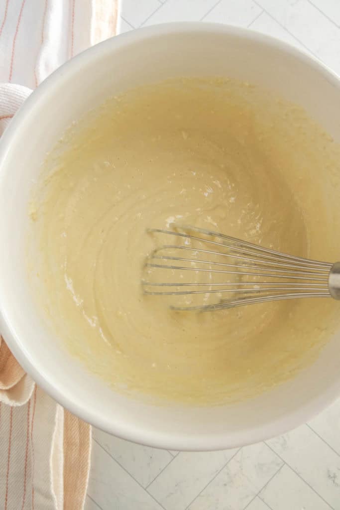 A metal whisk rests in a white mixing bowl filled with smooth, light yellow batter on a light-colored countertop.