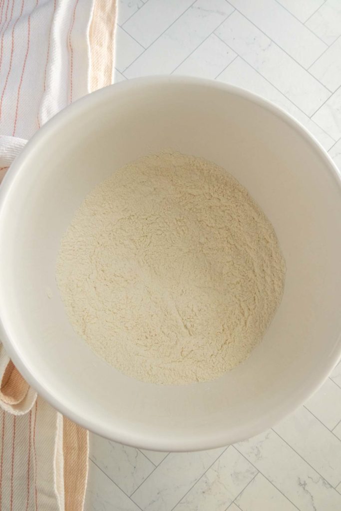 A white mixing bowl containing a mound of flour sits on a light-colored tiled surface next to a folded kitchen towel.
