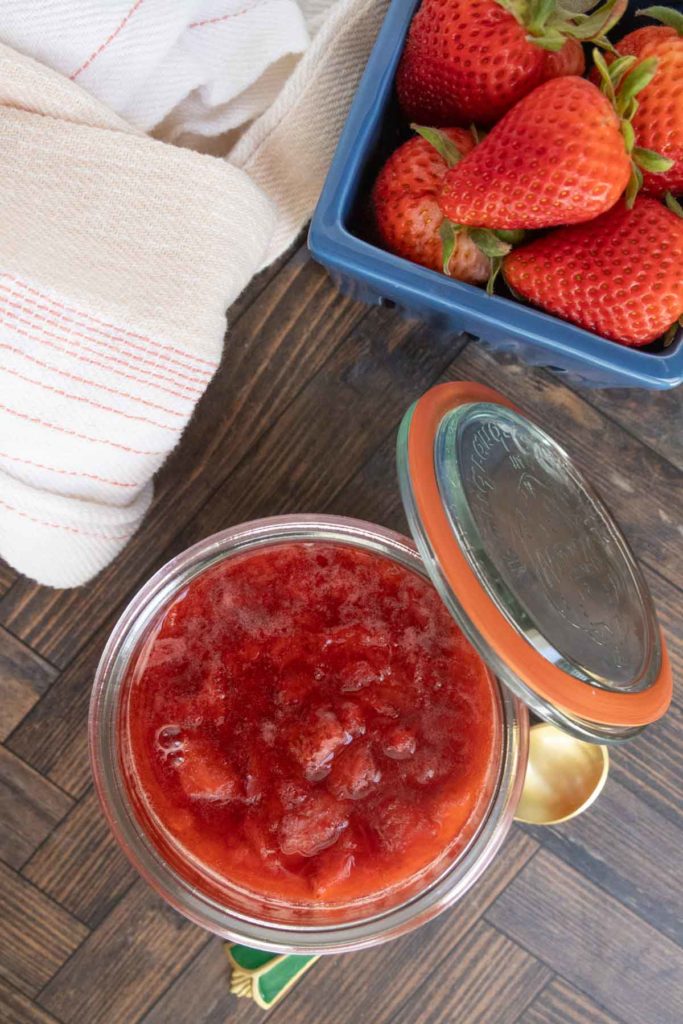 Open jar of strawberry jam placed next to a blue container of fresh strawberries and a white kitchen towel on a wooden surface.