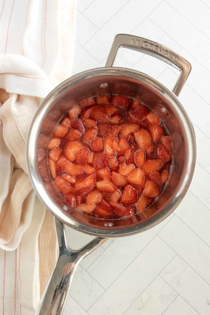 A stainless steel pot containing chopped strawberries simmering in liquid, placed on a light-colored countertop next to a folded towel.
