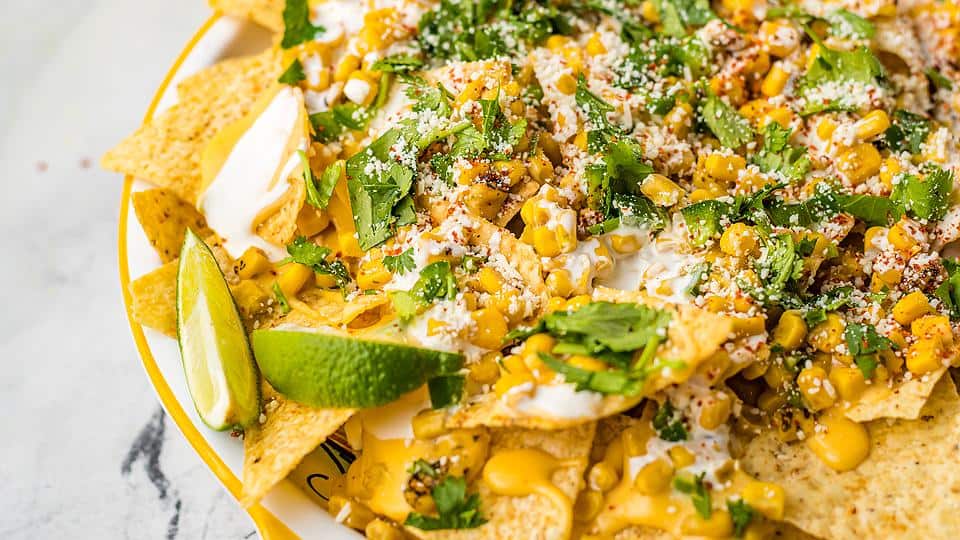 A plate of nachos topped with corn, cheese sauce, sour cream, cilantro, cotija cheese, and two lime wedges on the side.