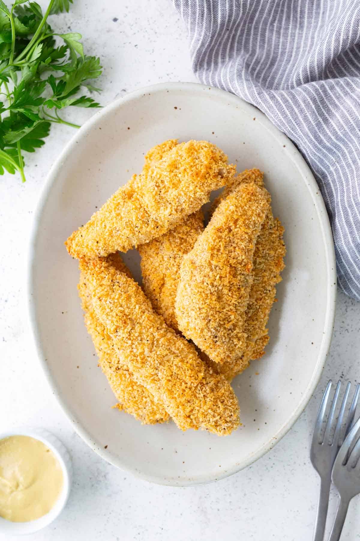 Four breaded chicken tenders on a white oval plate, with a sprig of parsley, a striped cloth, a small bowl of mustard, and two forks nearby.