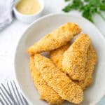 A plate of breaded chicken tenders is shown with a small dish of dipping sauce and fresh parsley in the background. Two forks are placed beside the plate.