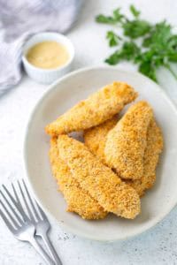 A plate of breaded chicken tenders is shown with a small dish of dipping sauce and fresh parsley in the background. Two forks are placed beside the plate.