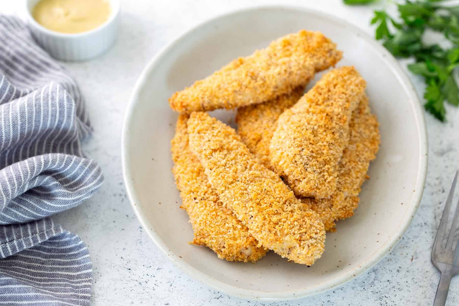 A white plate with several pieces of breaded baked chicken tenders, next to a striped napkin, parsley, and a small dish of dipping sauce.