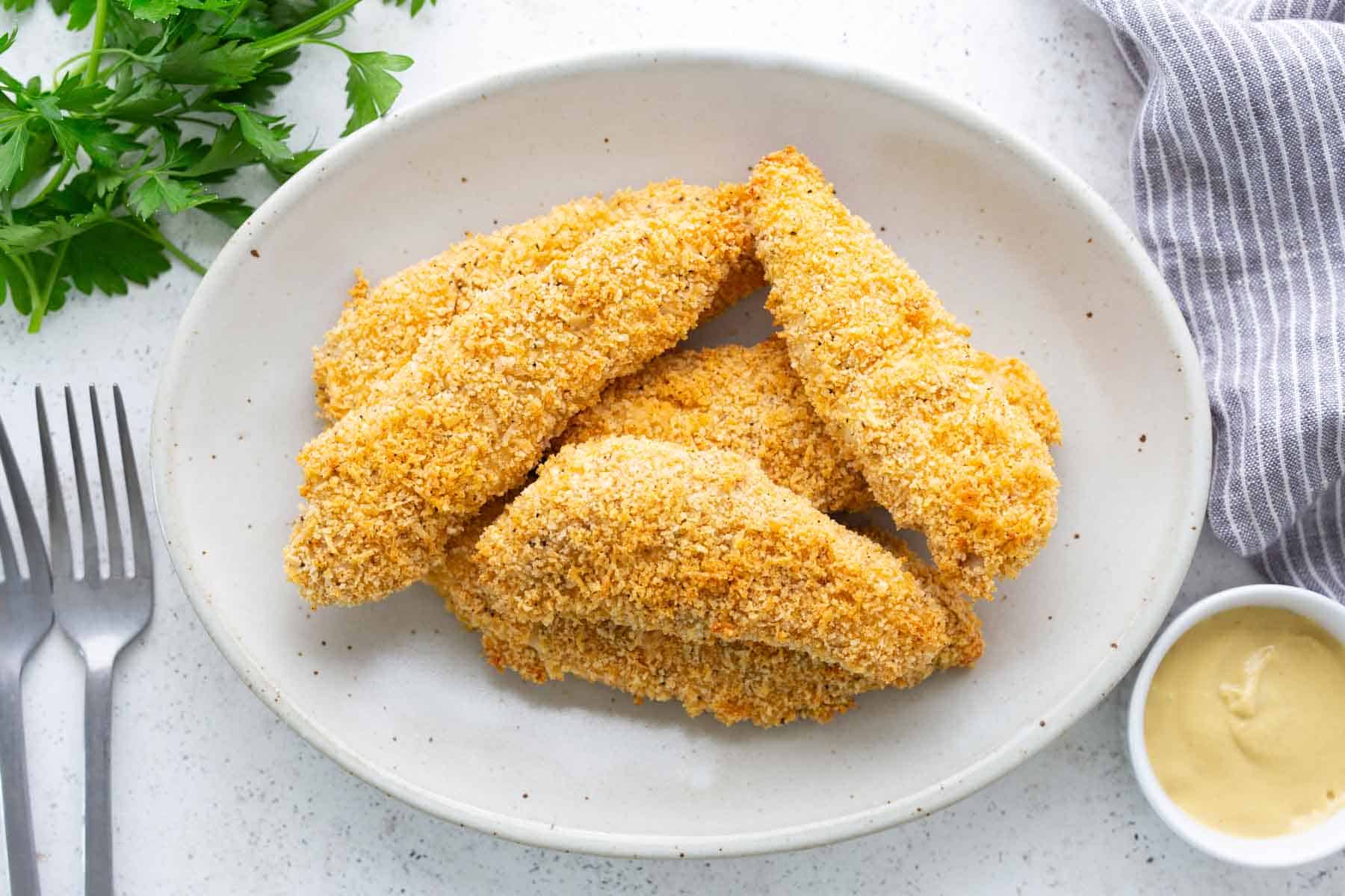 Four breaded chicken tenders on a white plate, served with a small bowl of dipping sauce, parsley, two forks, and a striped napkin nearby.