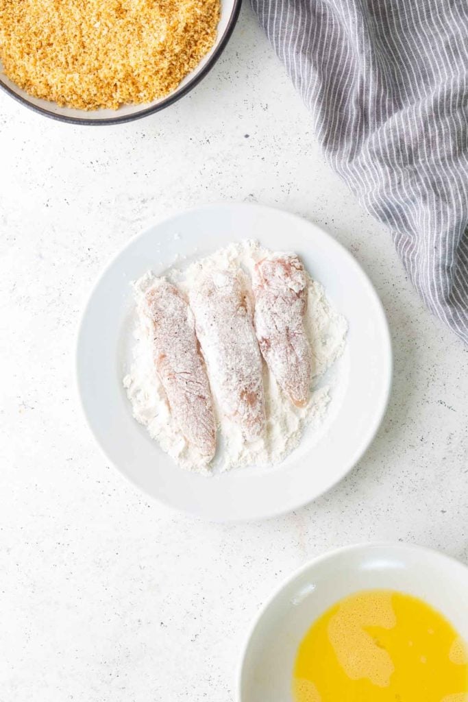 Three raw chicken tenders coated in flour on a white plate, with a bowl of beaten eggs and a bowl of breadcrumbs nearby. A gray striped cloth is visible in the upper right corner.