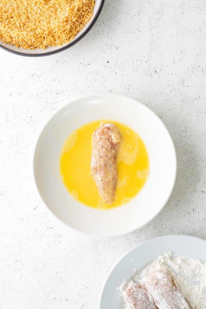 A piece of raw chicken is coated in egg wash in a white bowl, with bowls of breadcrumbs and flour nearby on a white surface.