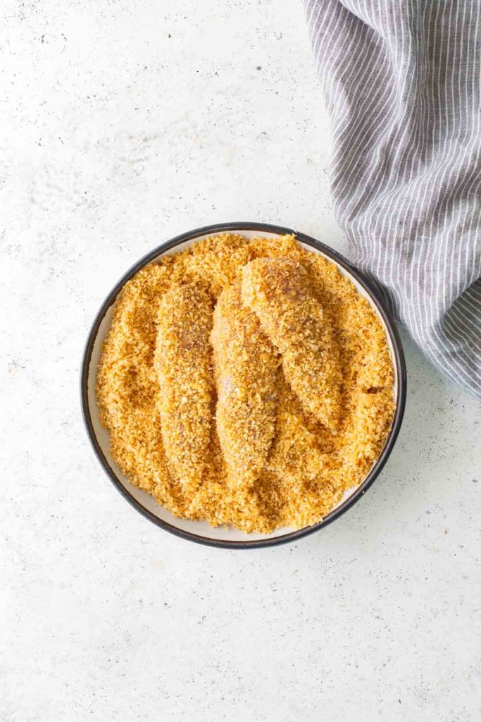 A bowl containing breaded chicken tenders on a light surface, with a striped cloth partially visible in the upper right corner.