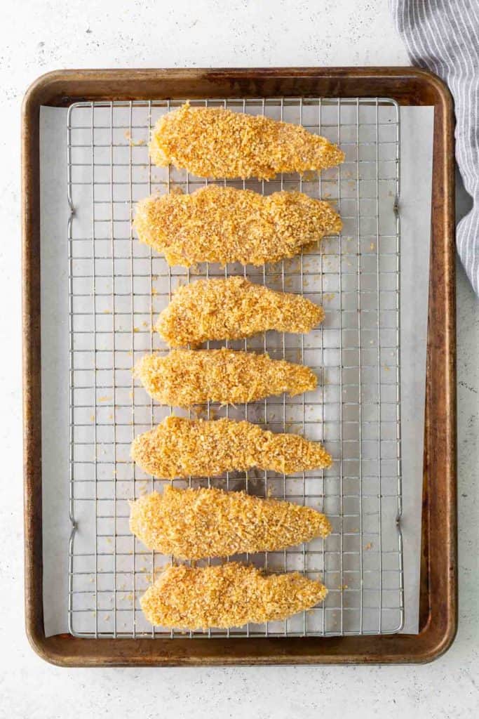 A baking sheet holds a wire rack with seven breaded chicken tenders arranged in a row, ready for baking.