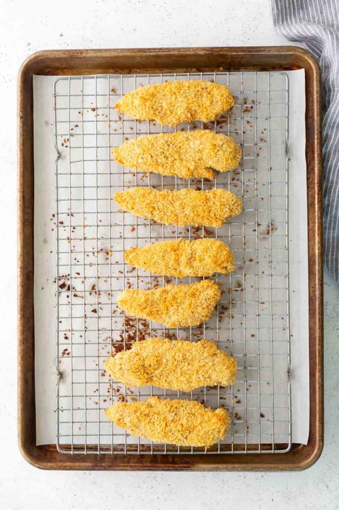 Six breaded chicken tenders arranged in a line on a wire rack over a baking sheet, ready to be cooked.