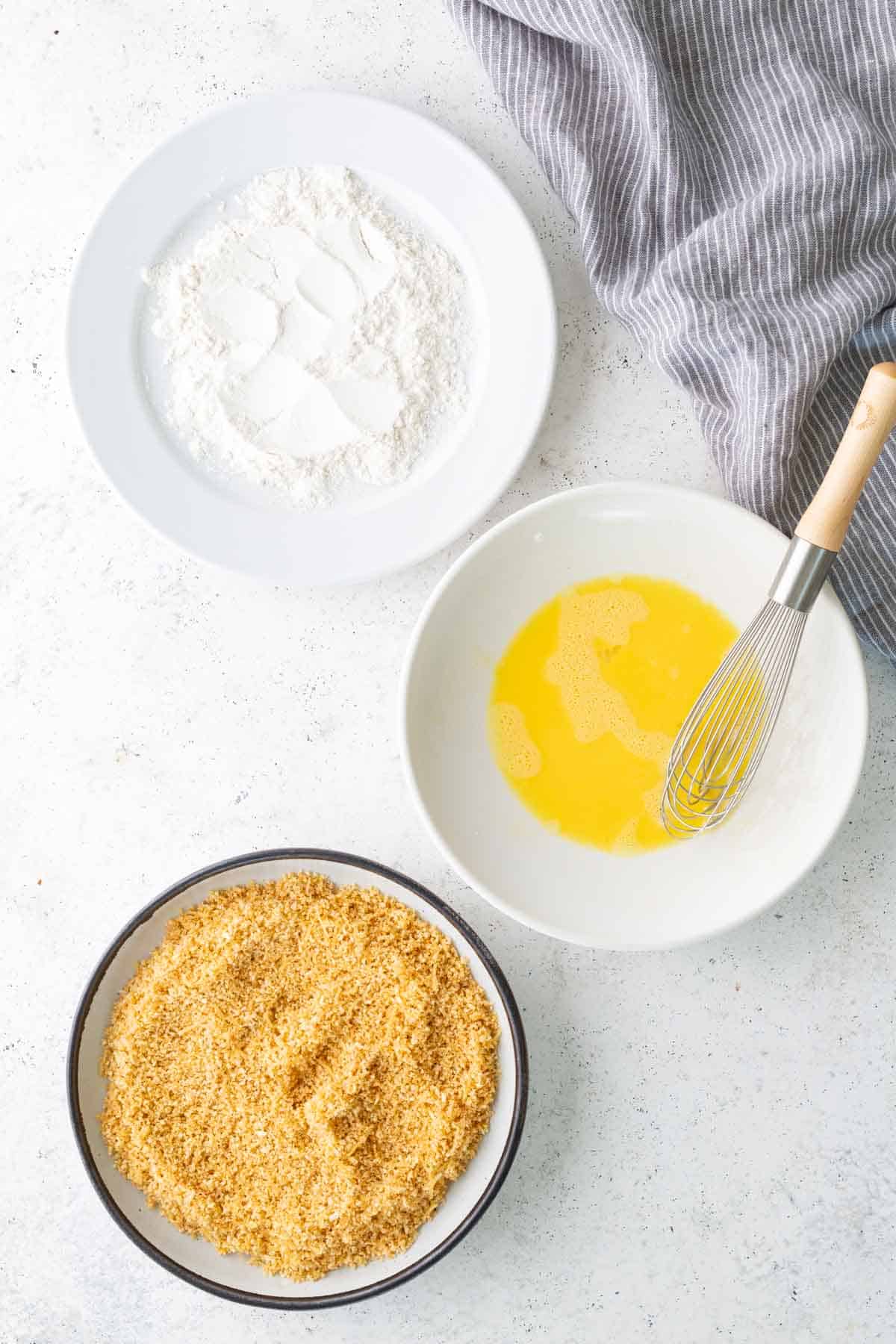 Three bowls on a white surface: one with flour, one with beaten eggs and a whisk, and one with breadcrumbs. A gray striped cloth is partially visible in the corner.