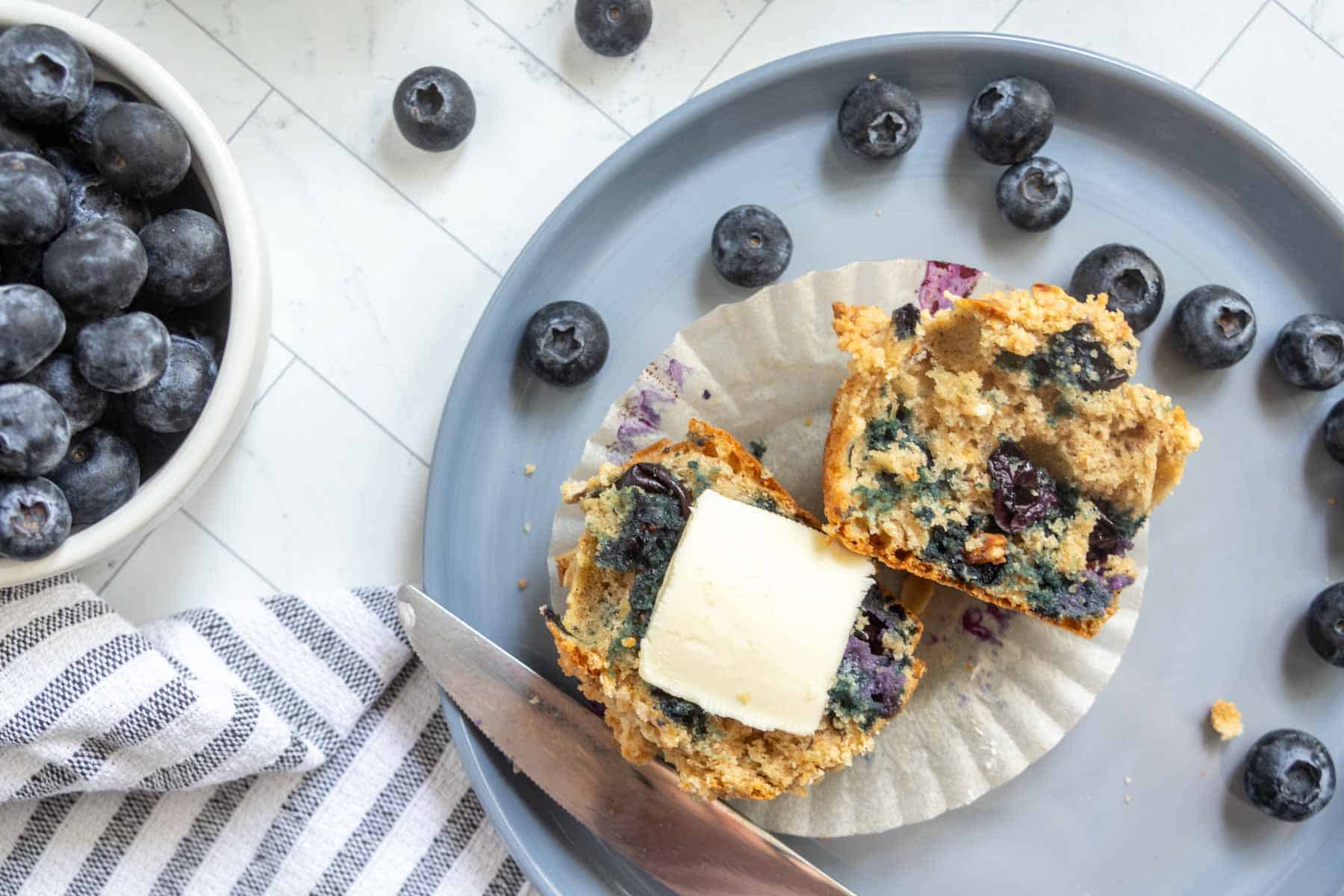 A split blueberry muffin with a pat of butter on a gray plate, surrounded by fresh blueberries, next to a bowl of blueberries and a striped napkin.
