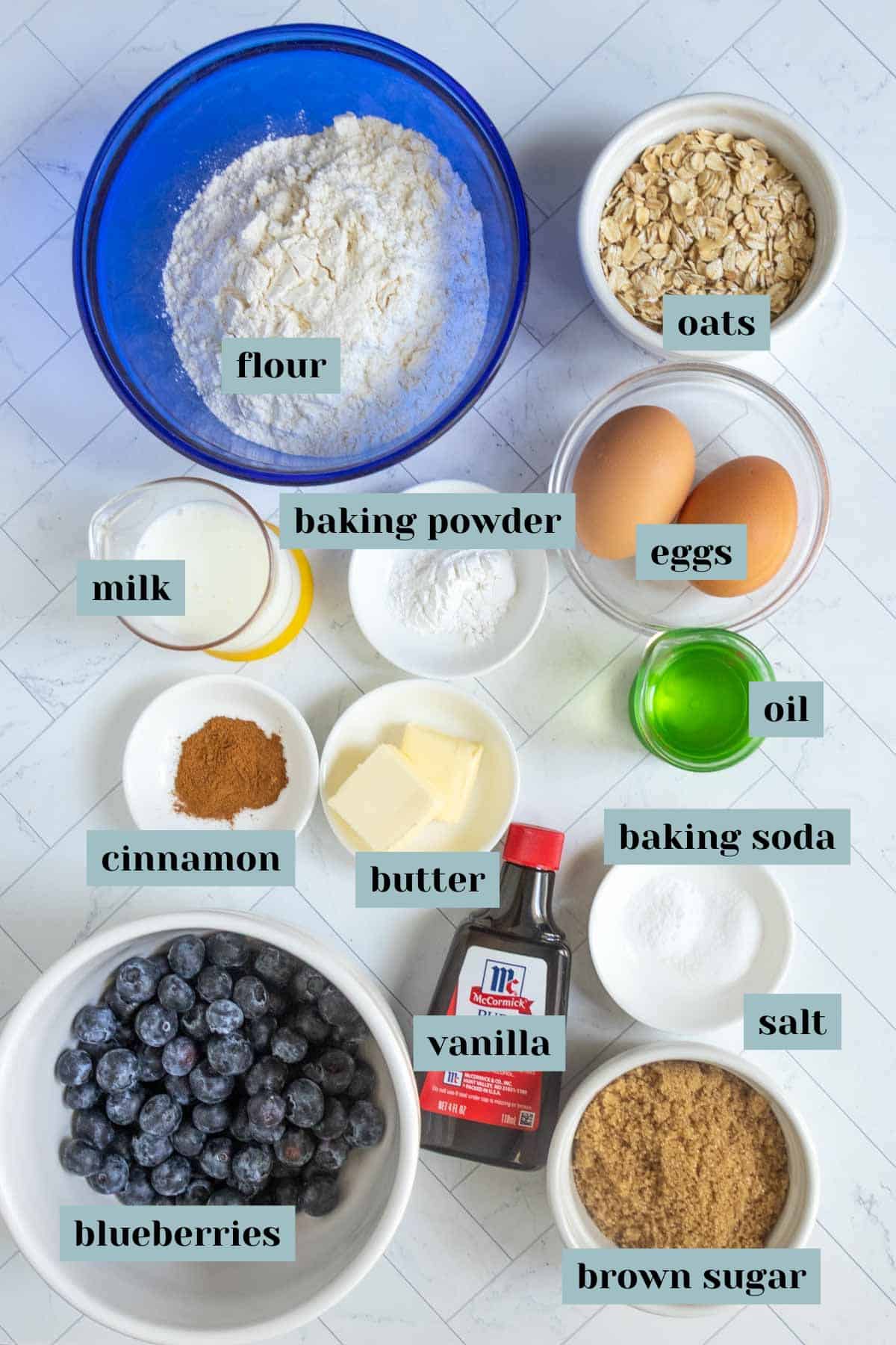 A variety of labeled ingredients for blueberry muffins are arranged on a white surface, including flour, oats, eggs, milk, oil, butter, spices, sugars, vanilla, salt, baking powder, and blueberries.