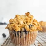 A blueberry muffin with a crumbly oat topping sits on a cooling rack.