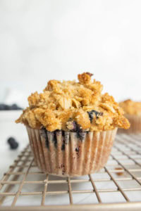 A blueberry muffin with a crumbly oat topping sits on a cooling rack.