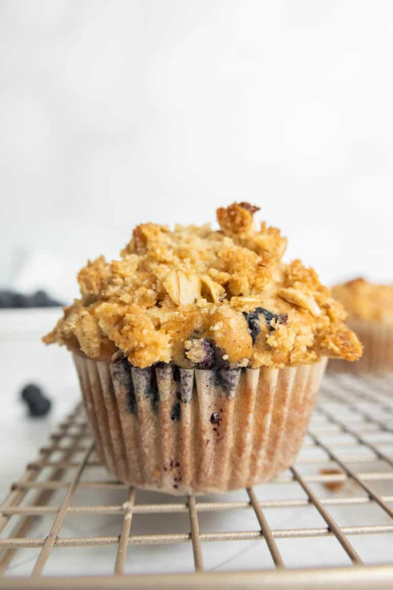 A blueberry muffin with a crumbly oat topping sits on a cooling rack.