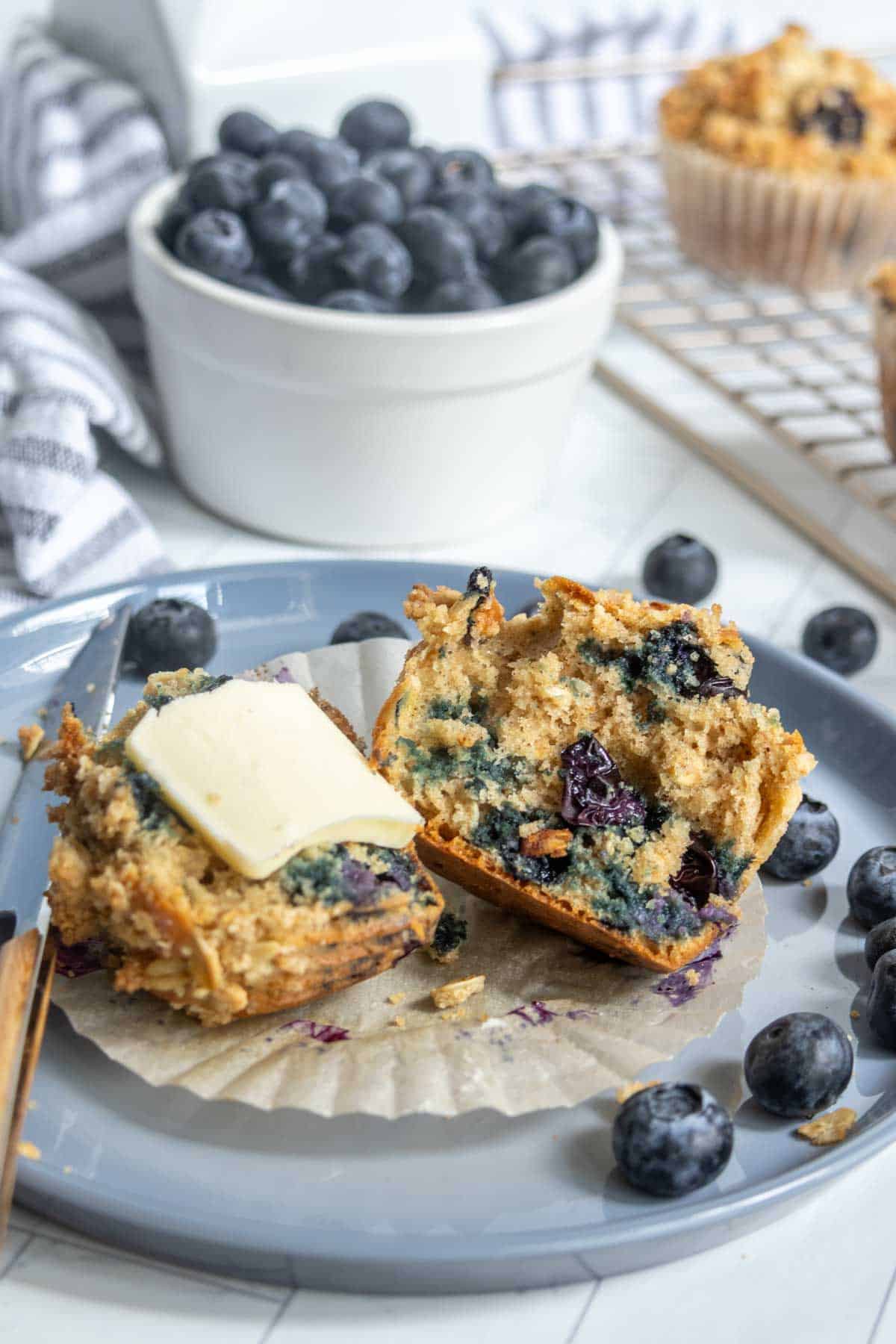 A blueberry muffin split in half on a plate, one half topped with a slice of butter, surrounded by fresh blueberries.