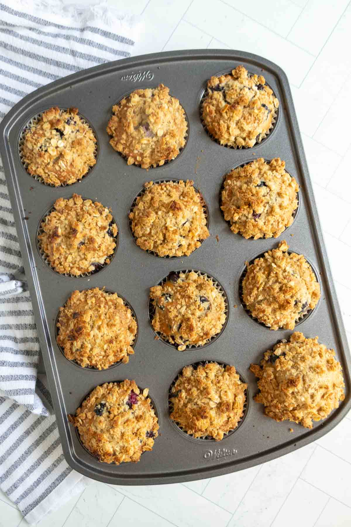 A muffin tin with twelve baked oatmeal muffins, some containing visible blueberries, sits on a striped kitchen towel on a light-colored surface.
