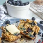 A blueberry streusel muffin split in half on a plate, one half topped with butter; fresh blueberries and a bowl of blueberries in the background.