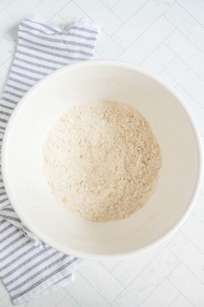 A white mixing bowl filled with dry flour mixture sits on a white tiled surface next to a gray and white striped cloth.