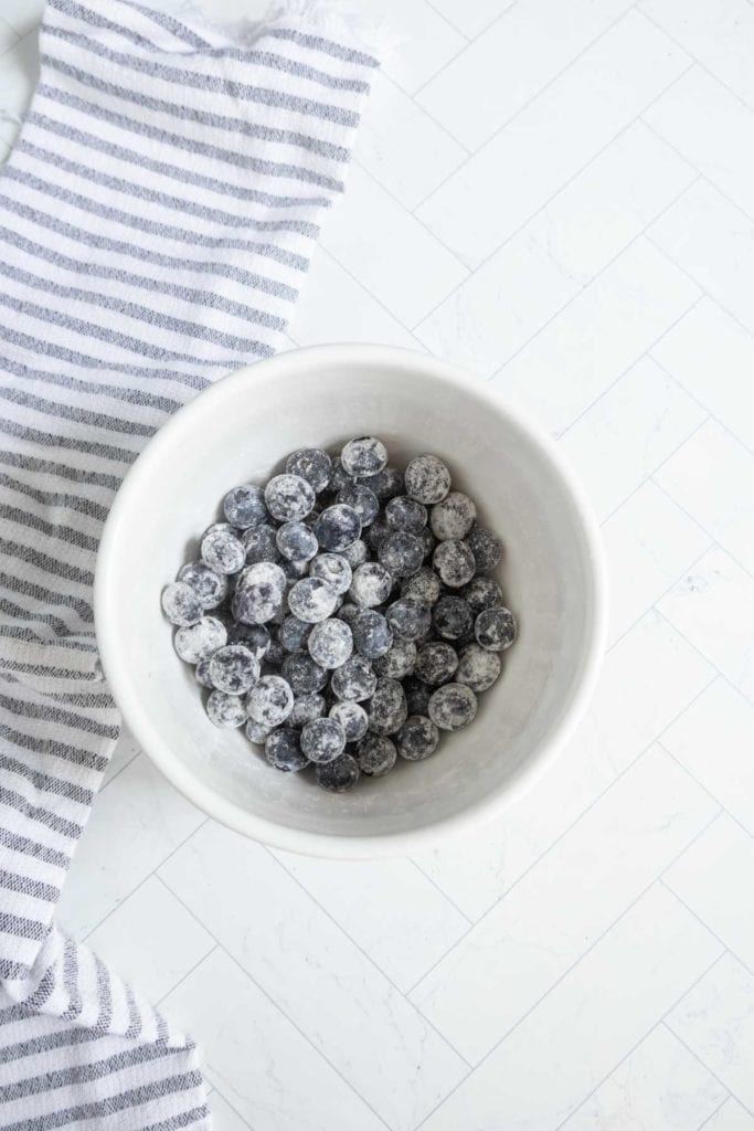 A white bowl filled with blueberries coated in flour, placed on a white tiled surface next to a gray and white striped cloth.