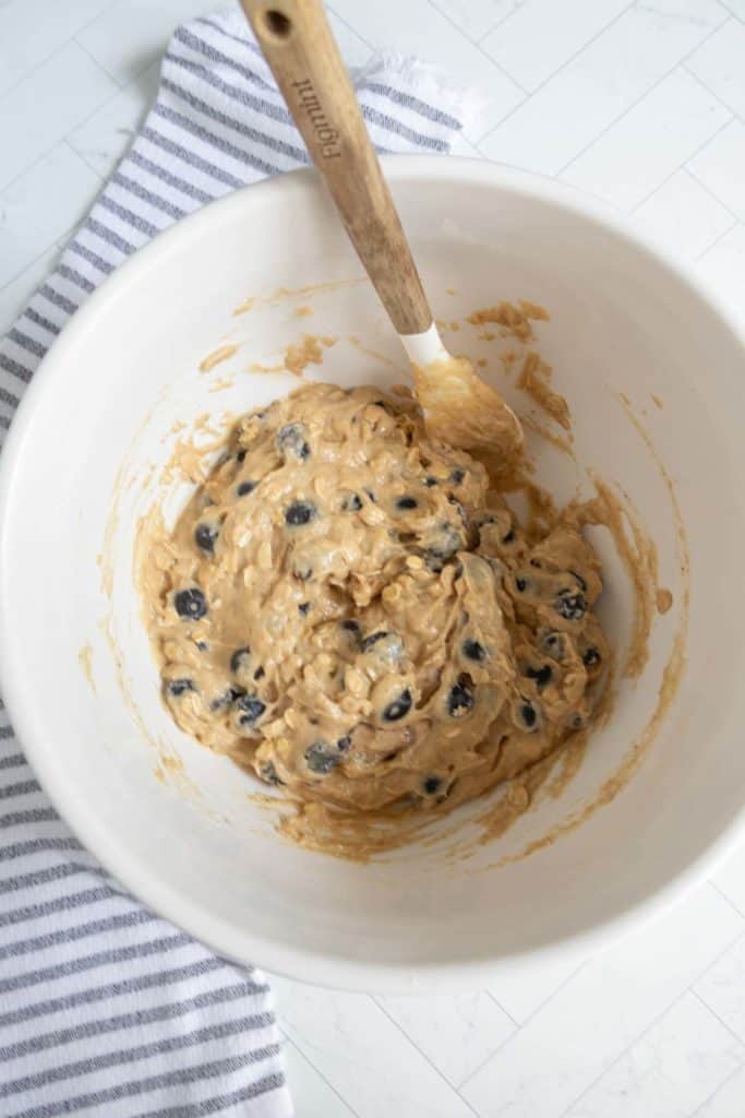 A white mixing bowl with a wooden spatula contains cookie dough with chocolate chips on a white surface next to a striped towel.