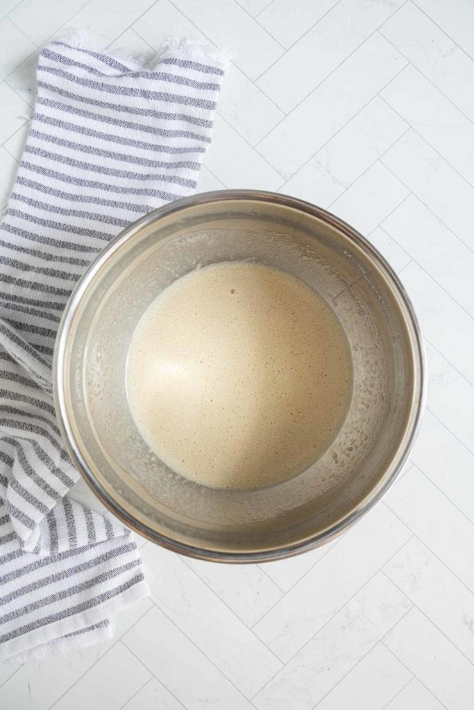 A metal mixing bowl filled with a light, creamy batter, placed on a white tiled surface next to a gray and white striped kitchen towel.