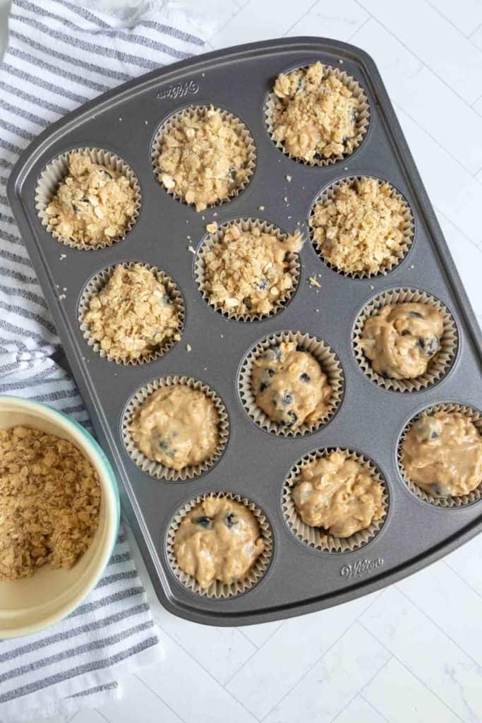 A muffin tin filled with twelve paper liners containing unbaked muffin batter and crumb topping; some muffins have more topping than others. A bowl with extra topping sits nearby.