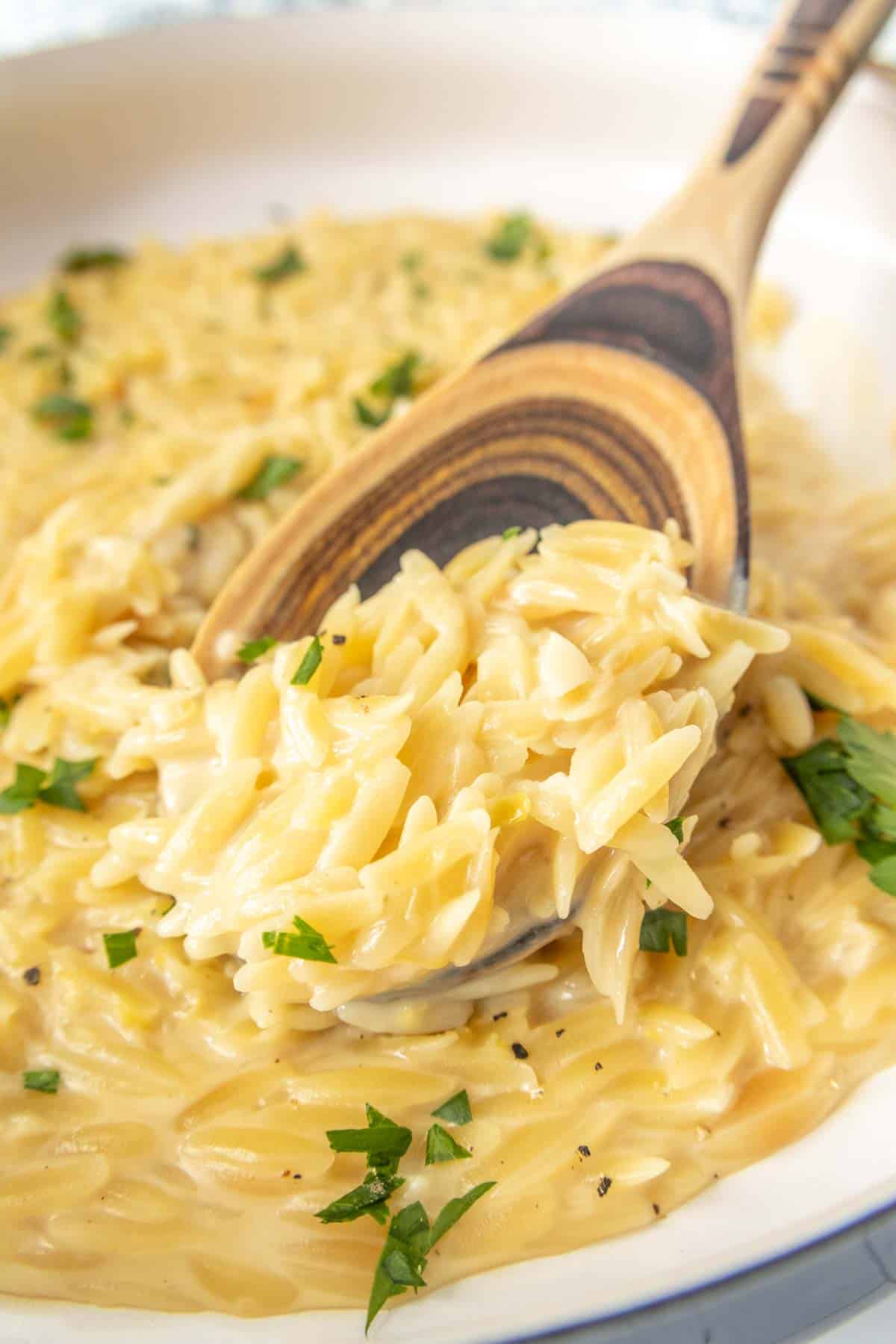 A wooden spoon scooping creamy lemon orzo pasta, garnished with chopped parsley and black pepper, from a white bowl.