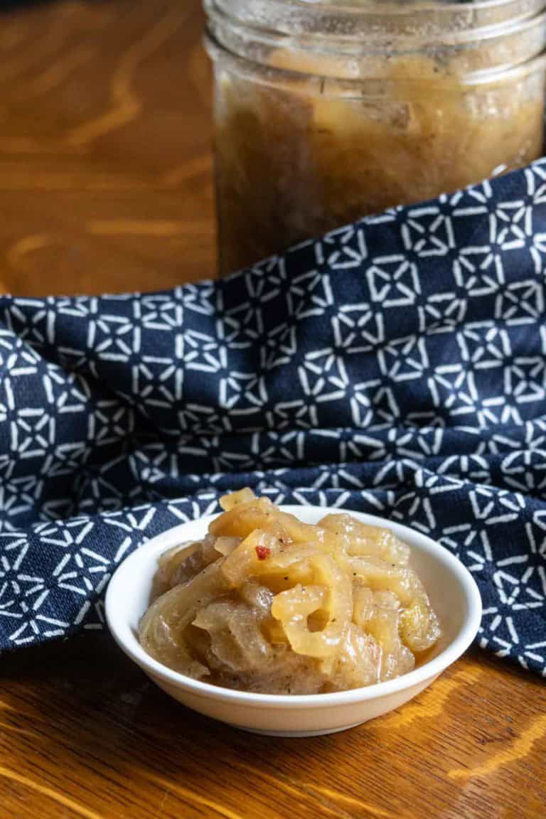 A small white bowl filled with caramelized onions sits in front of a jar, with a patterned blue and white cloth underneath on a wooden surface.