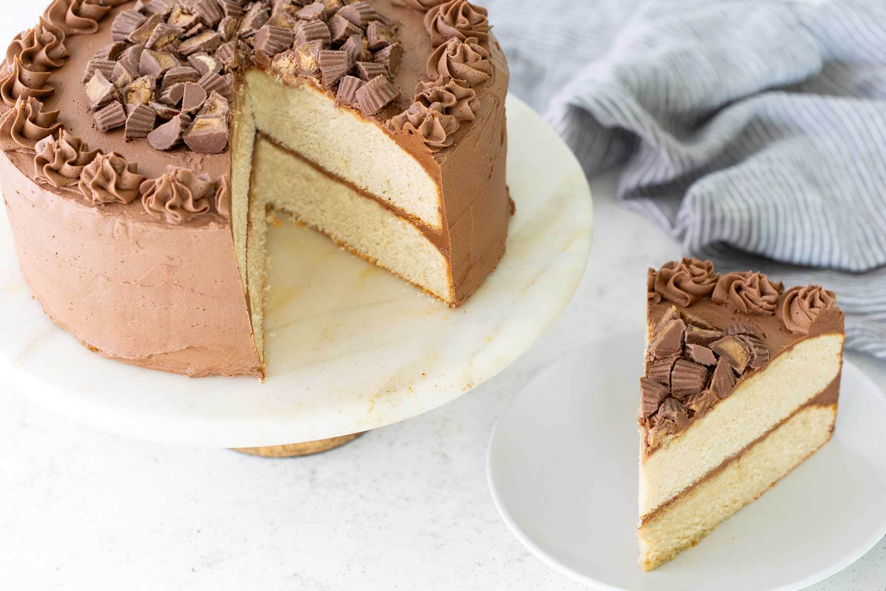 A round layer cake with chocolate frosting and chopped peanut butter cups on top, sitting on a marble cake stand, with one slice served on a white plate.