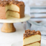 A slice of two-layer vanilla cake with chocolate frosting sits on a white plate; the rest of the cake is on a cake stand in the background.