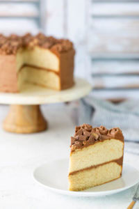 A slice of two-layer vanilla cake with chocolate frosting sits on a white plate; the rest of the cake is on a cake stand in the background.